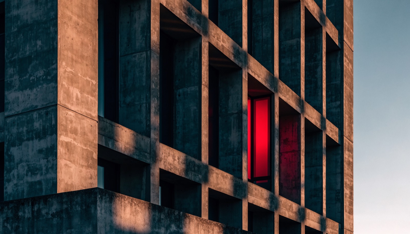 A concrete facade at golden hour with deep recessed windows, one lit crimson from inside.
